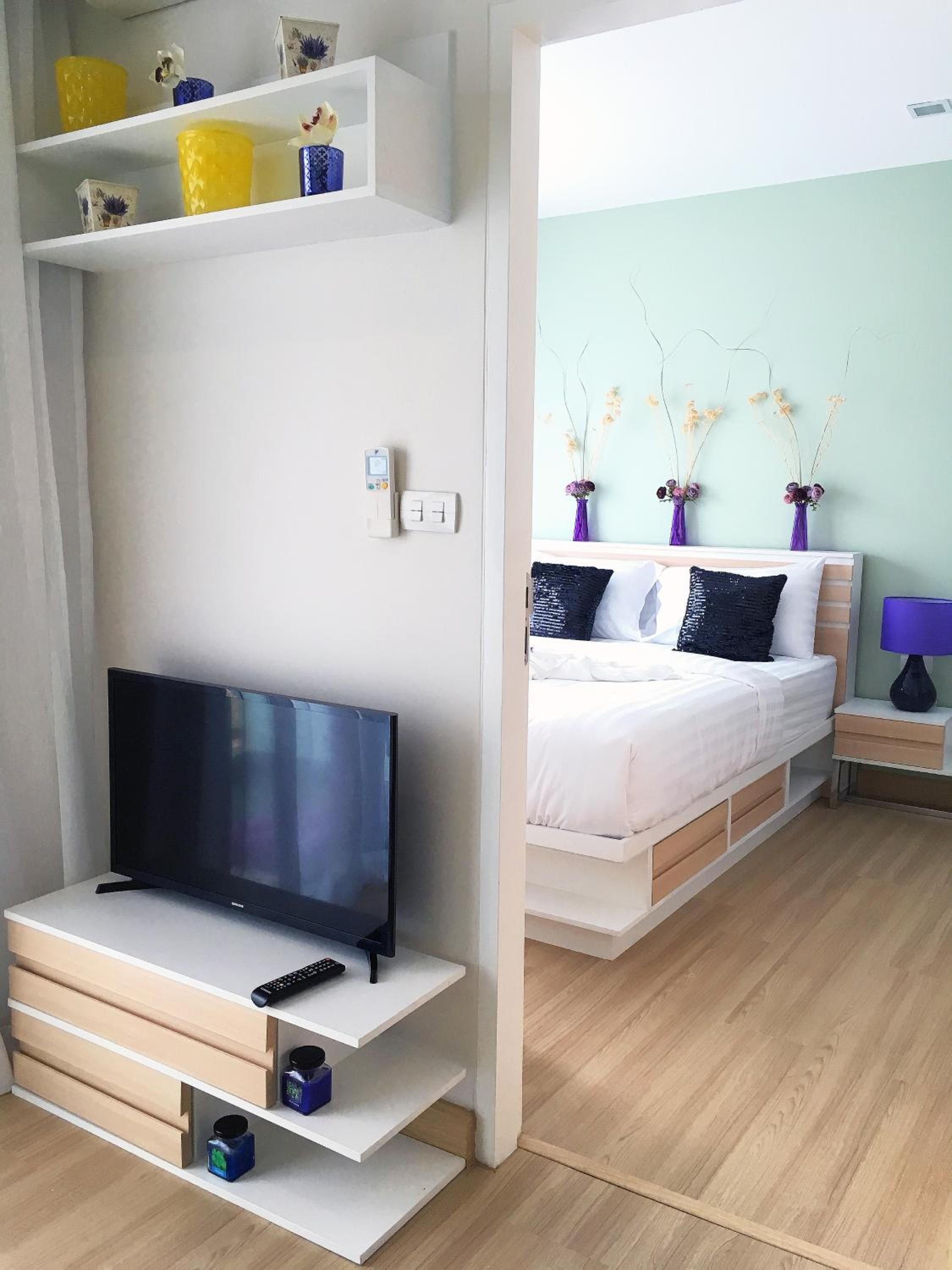 A modern bedroom with a flat-screen TV, a bed with white bedding, and a bedside table with a lamp.