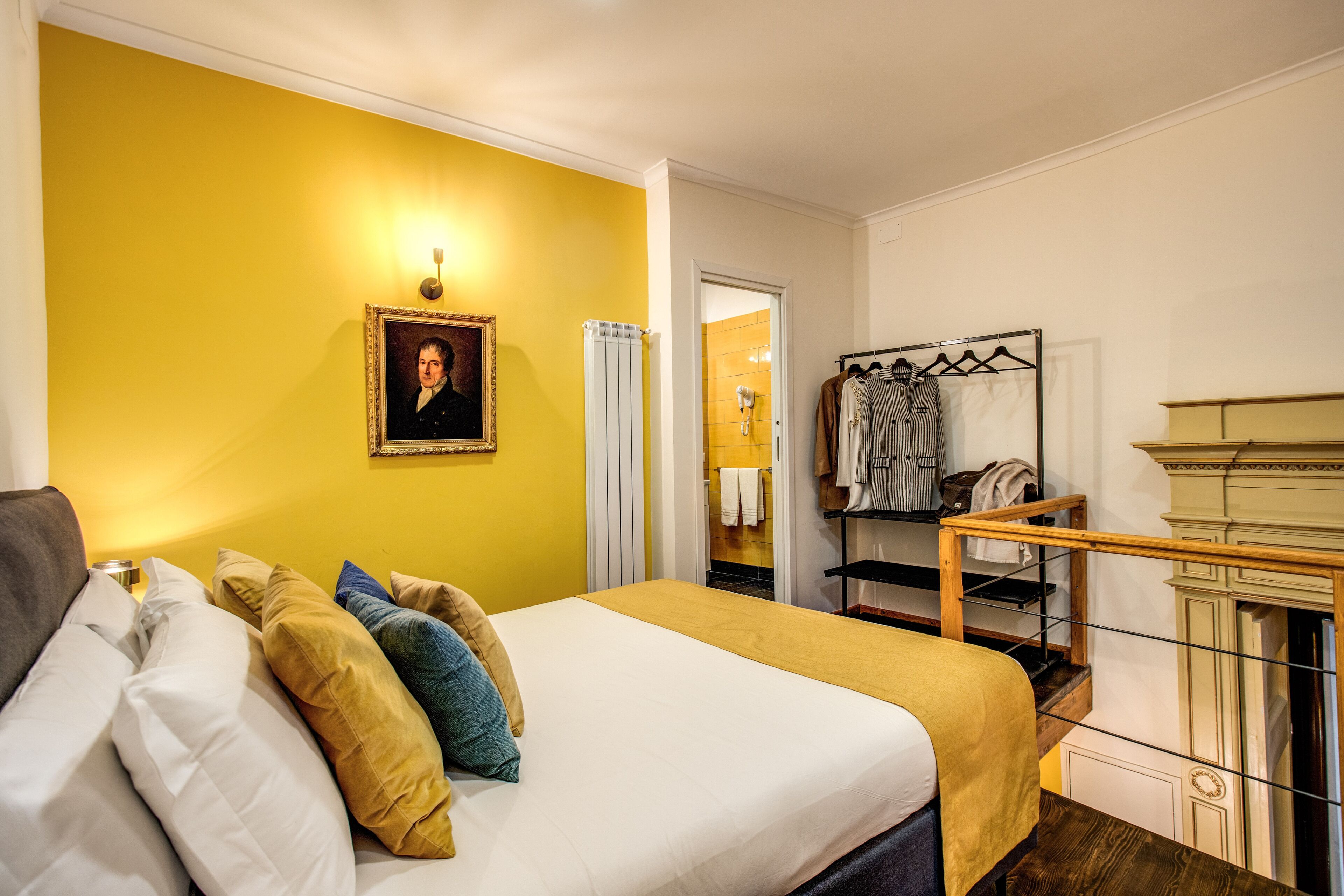 A bedroom with a bed, pillows, a framed portrait, a radiator, a clothing rack, and a staircase.