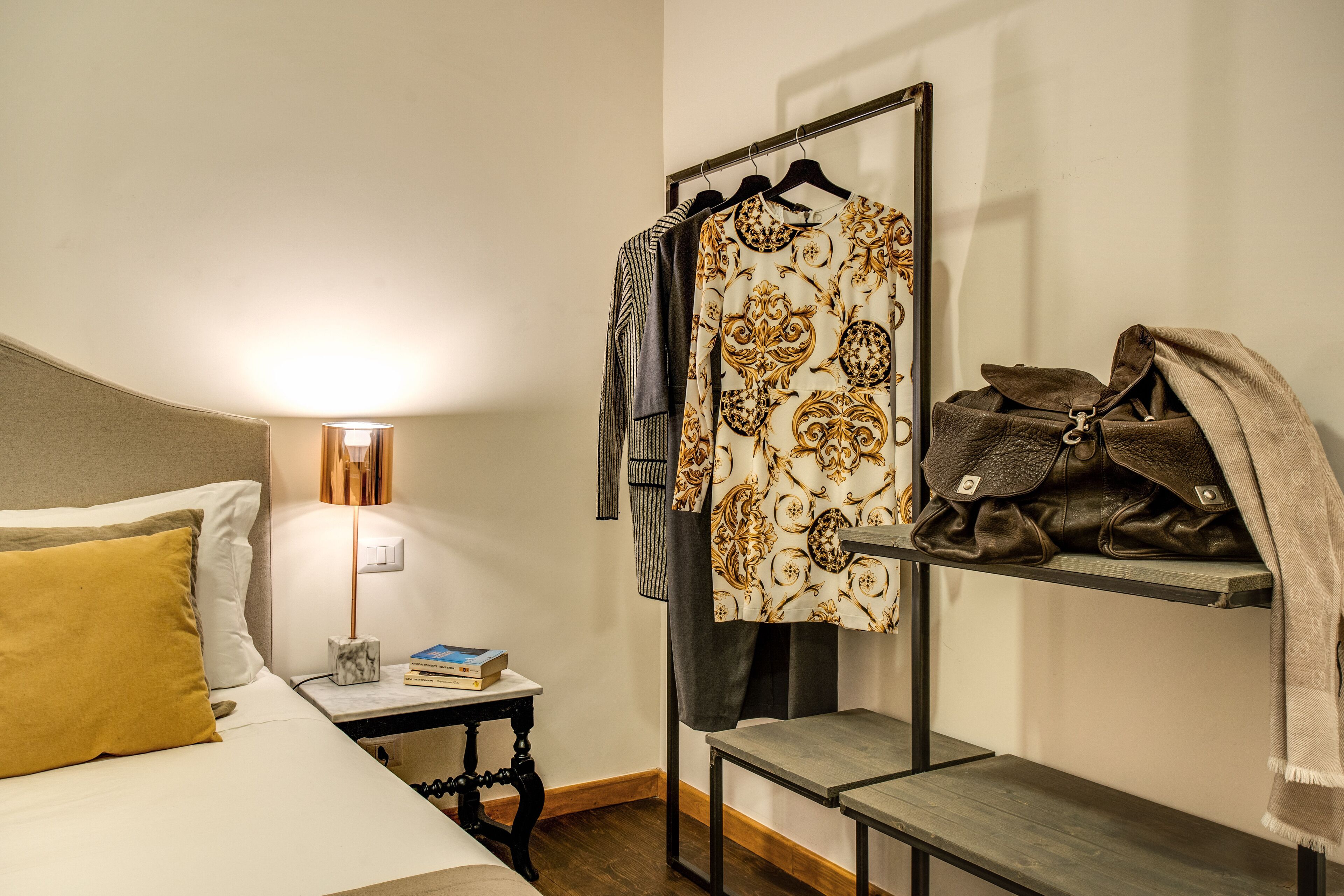 Habitación de hotel ordenada con cama, mesita de noche, lámpara y un perchero con una prenda estampada y un bolso de cuero.