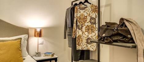 Habitación de hotel ordenada con cama, mesita de noche, lámpara y un perchero con una prenda estampada y un bolso de cuero.