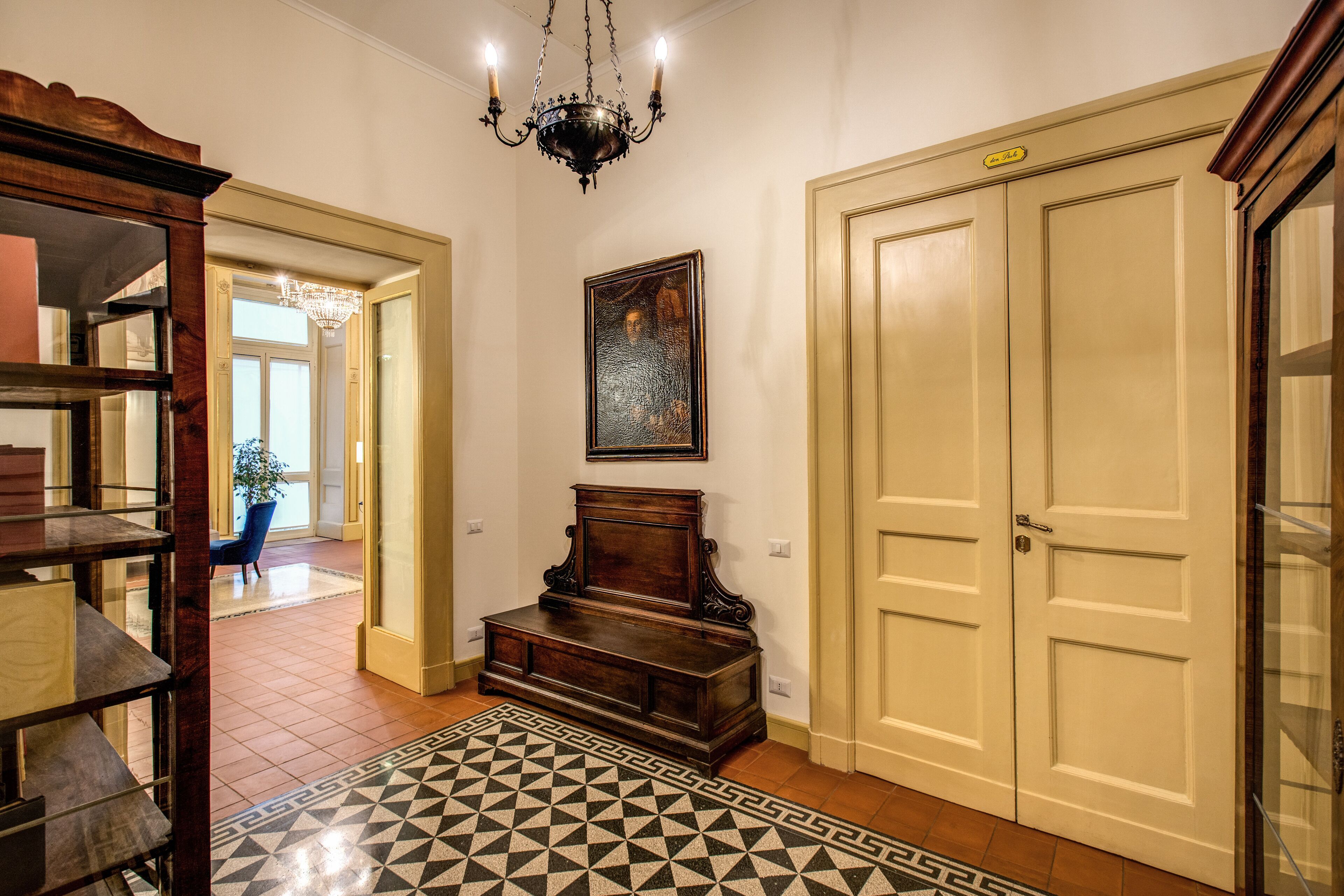 A hallway with a patterned rug, a wooden bench, a chandelier, and a door with a sign above it.