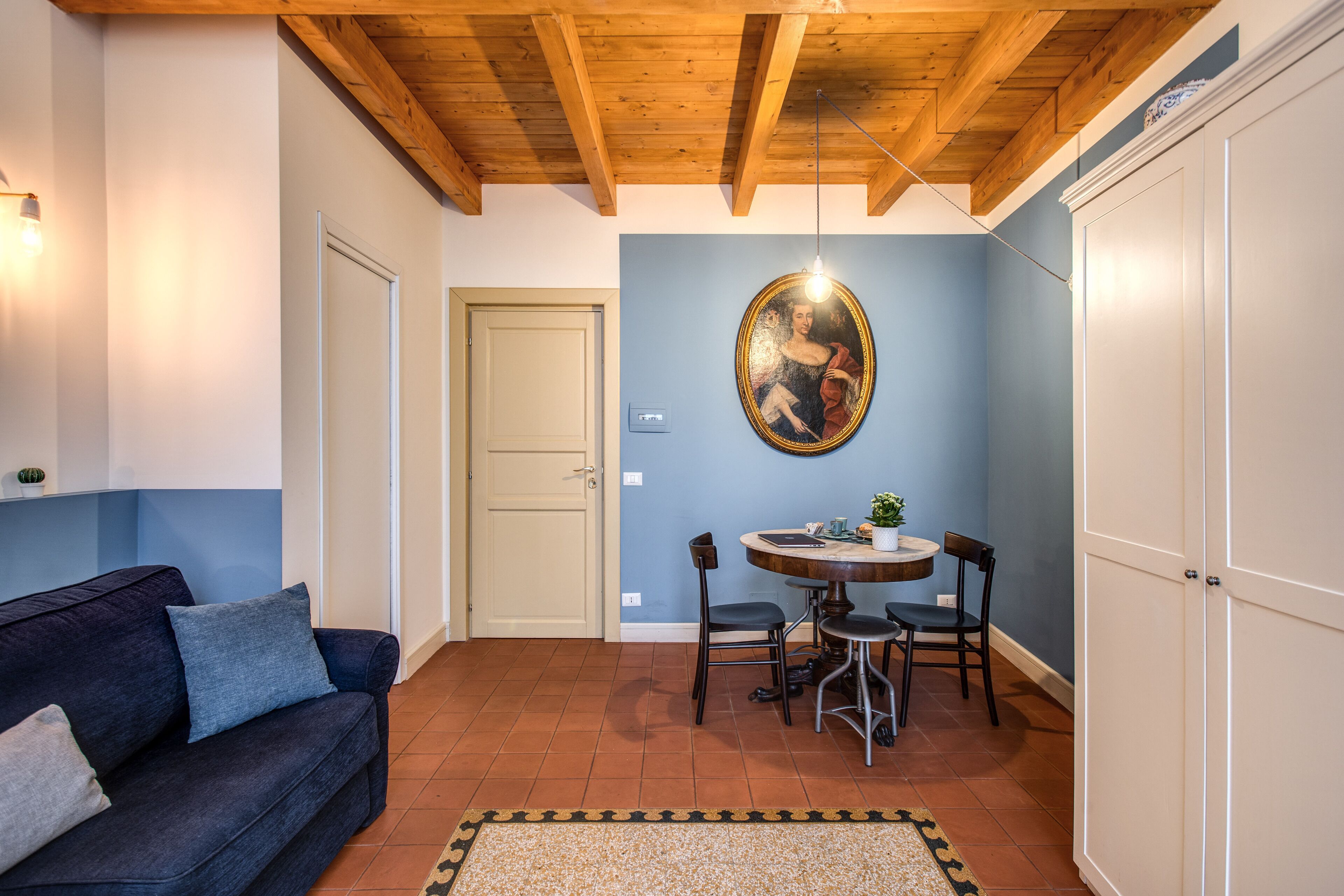 Un salon avec un canapé bleu foncé, une table de salle à manger ronde entourée de chaises, un plafond en bois et une armoire blanche.