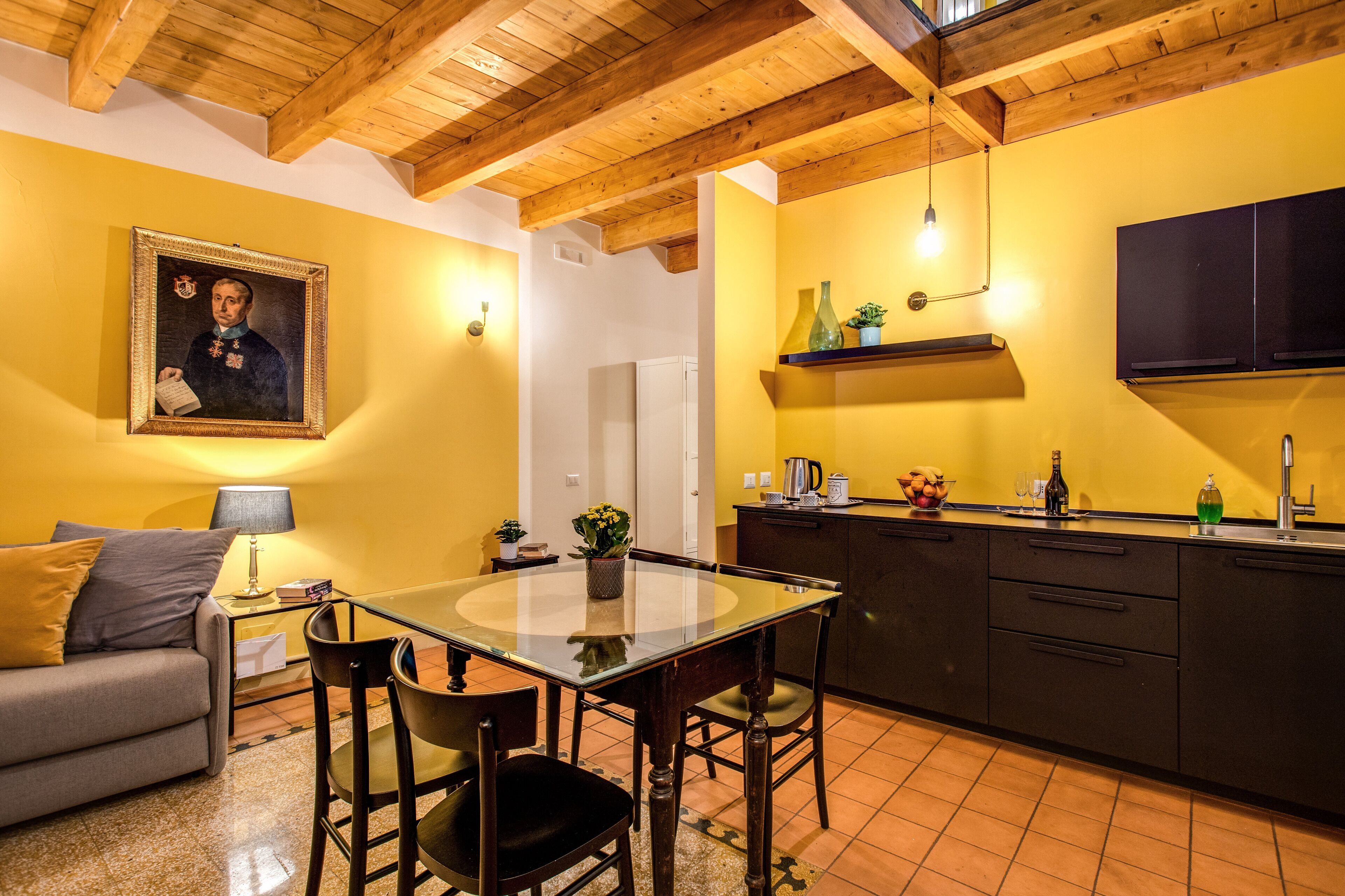 A dining area with a glass table, chairs, and a sofa. There is a painting on the wall, a lamp, and a kitchen area with cabinets and a sink.