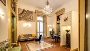 A spacious living room with a chandelier, a sofa with a yellow cushion, a dining table with chairs, and a decorative floor rug.