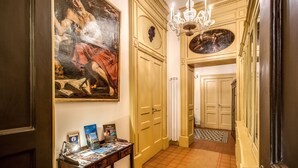 A hallway with a large classical painting, a chandelier, and a table with brochures.