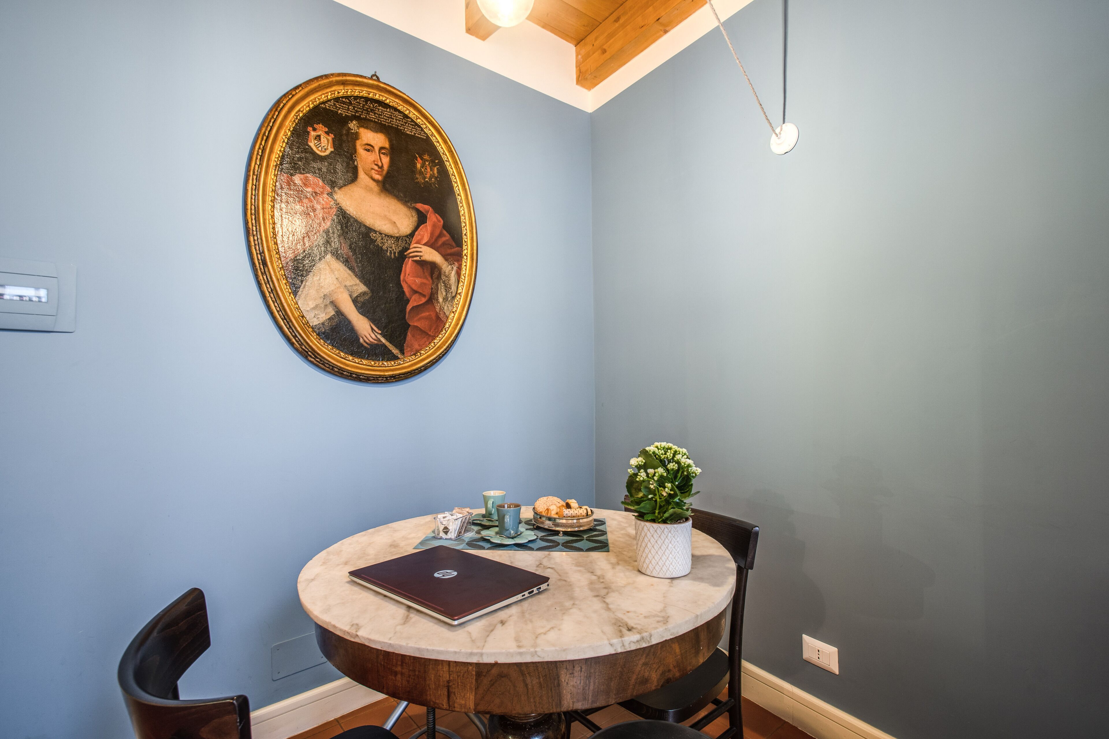A dining area with a round table, chairs, a laptop, a potted plant, and a painting on the wall.