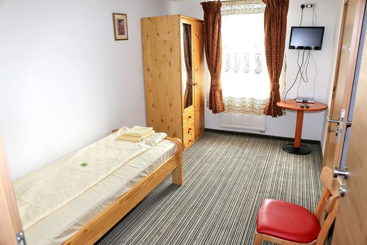 Een kleine, netjes ingerichte kamer met een enkel bed, een houten garderobe, een klein tafeltje met een televisie en een rode stoel.