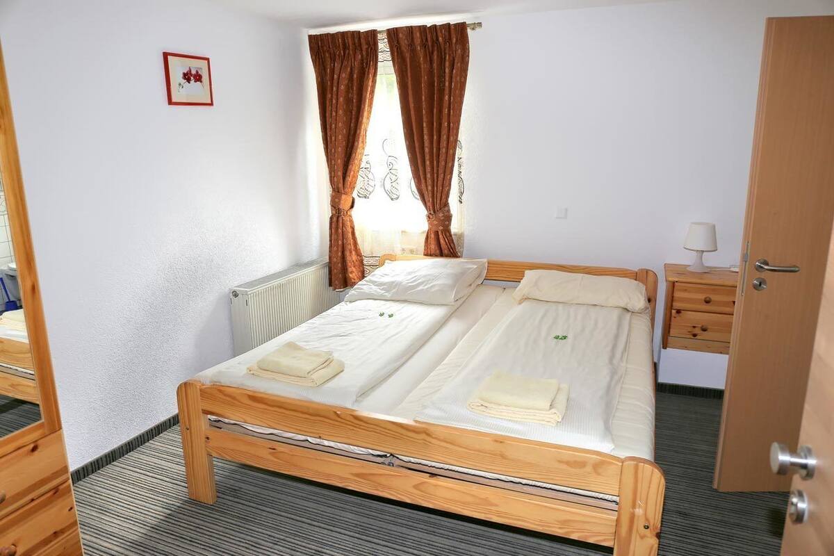 Een eenpersoonskamer met een houten bedframe, wit beddengoed, een radiator, een houten nachtkastje, een lamp en een deur.