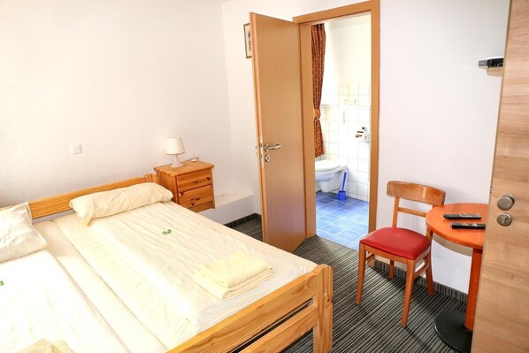 Una habitación pequeña y ordenada con una cama individual, una silla de madera, una mesita con un control remoto y una puerta que da a un baño.