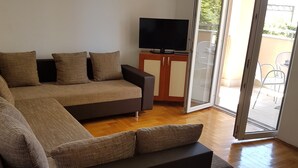Un salon moderne avec un canapé d’angle, un téléviseur à écran plat posé sur un meuble en bois et une porte-fenêtre coulissante donnant sur une terrasse extérieure.