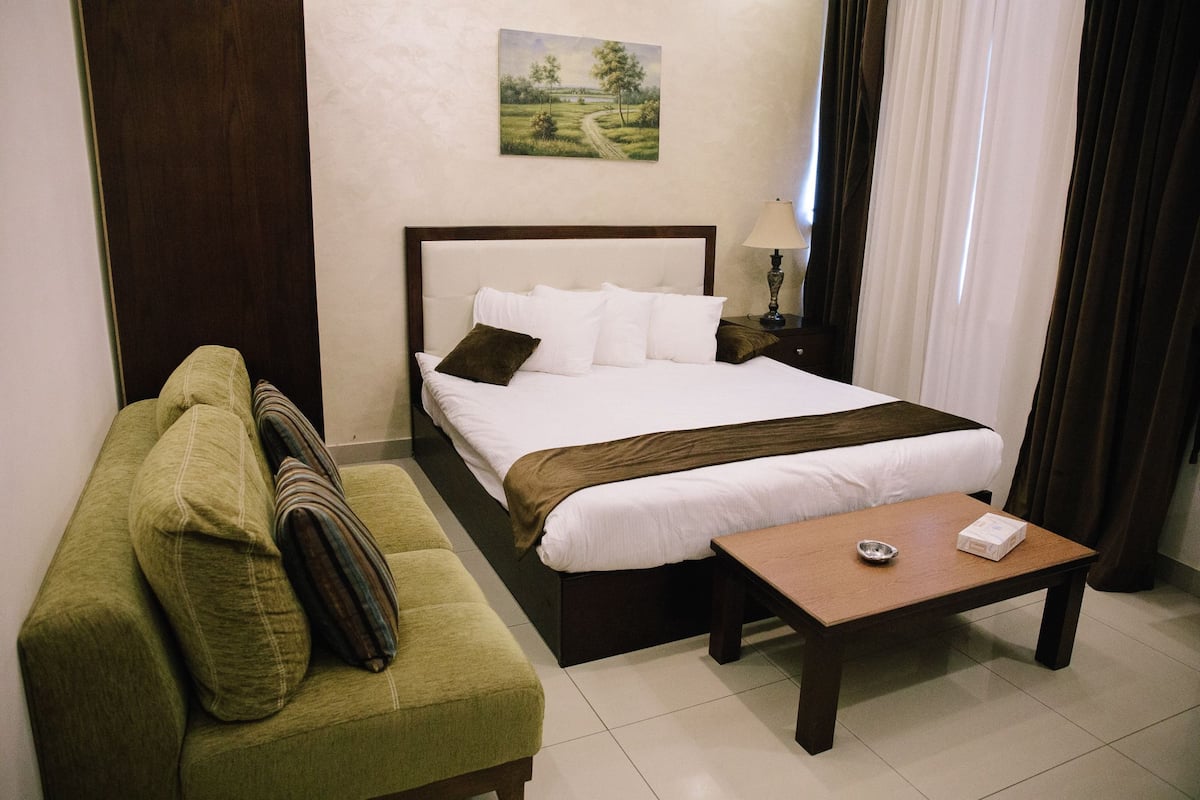 A neatly arranged hotel room with a bed, a green sofa, a wooden table, and a painting on the wall.