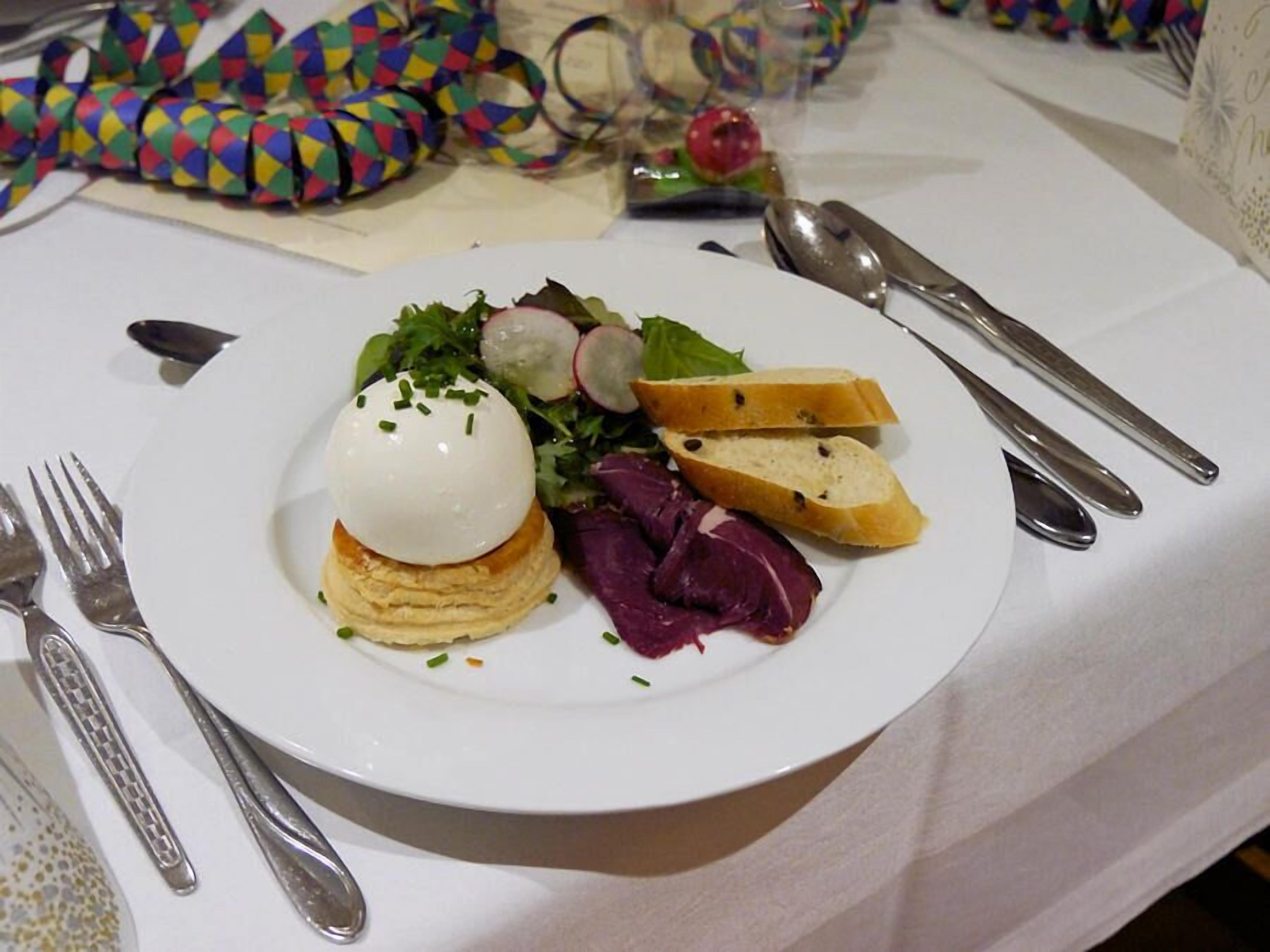 Un plato con un huevo escalfado sobre tostada, rodajas de rábano y una ensalada, acompañado por un tenedor y una cuchara sobre una mesa con mantel blanco.