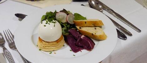 A plate with a poached egg on toast, radish slices, and a salad, accompanied by a fork and spoon on a table with a white tablecloth.