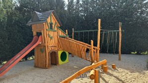 A wooden play structure with a slide and climbing elements, set on sandy ground with trees in the background. - Hotel Forsthaus Luchsenburg (Ohorn)