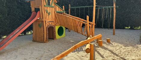 A wooden play structure with a slide and climbing elements, set on sandy ground with trees in the background.