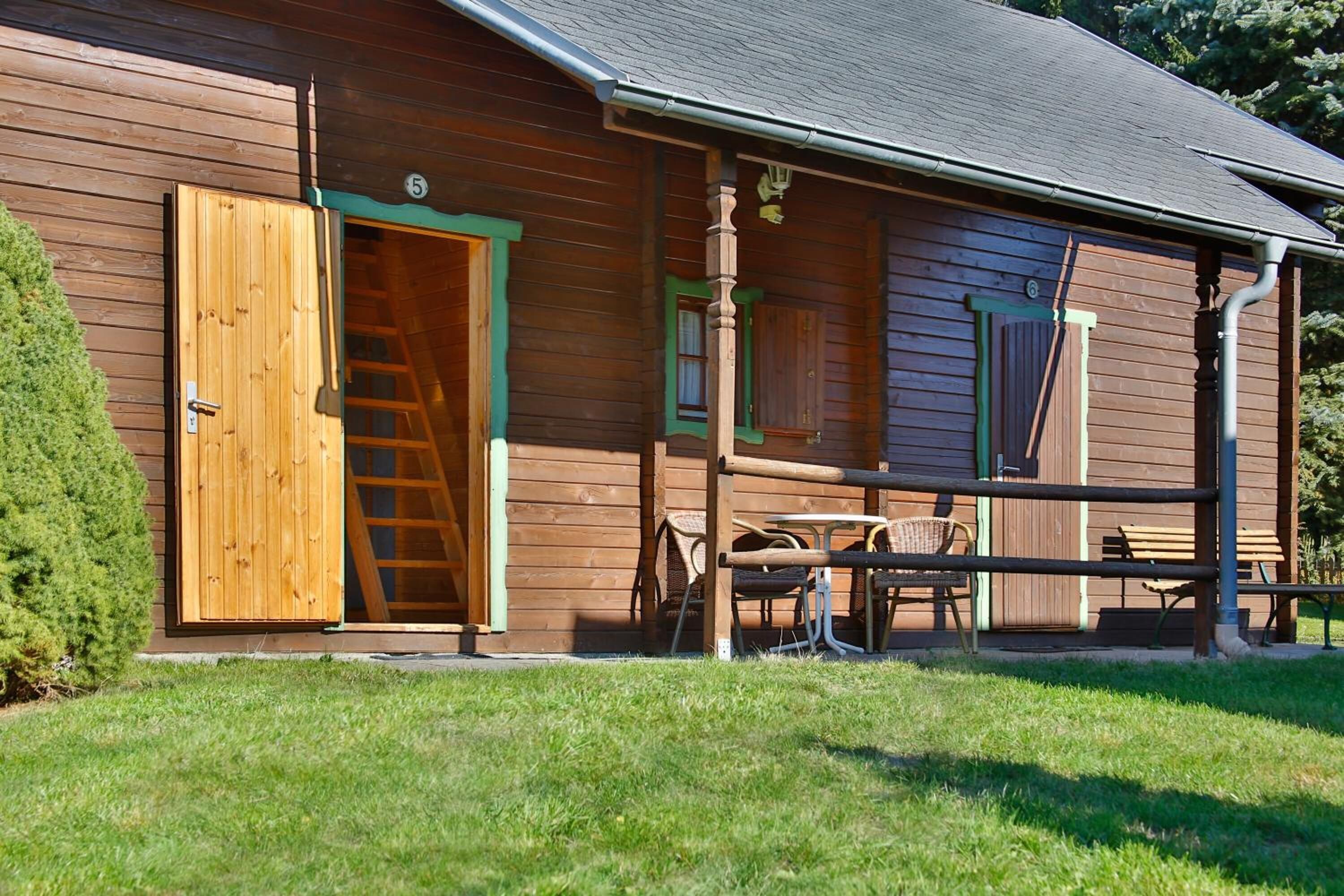 A wooden cabin with a porch, a wooden door, a ladder, and a table with chairs.
