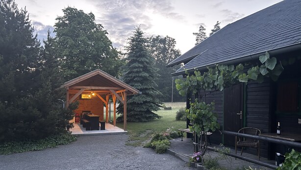 A covered outdoor seating area with a table and benches, a gravel path leading to a building with a dark roof, and greenery surrounding the space. - Hotel Forsthaus Luchsenburg (Ohorn)