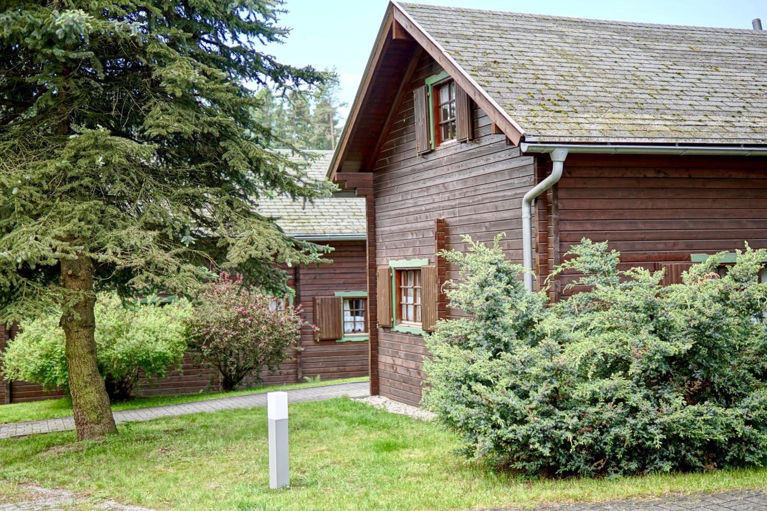 A wooden cabin with a sloped roof, surrounded by dense greenery and a paved pathway.