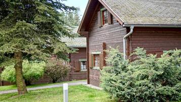 A wooden cabin with a sloped roof, surrounded by dense greenery and a paved pathway.