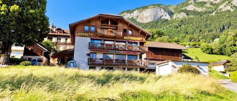 A hotel with a restaurant on the second floor, surrounded by lush greenery and mountains.
