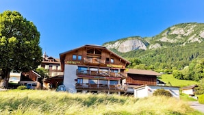 A hotel with a restaurant on the second floor, surrounded by lush greenery and mountains. - Logis Hotel L'accueil Savoyard (Mieussy)