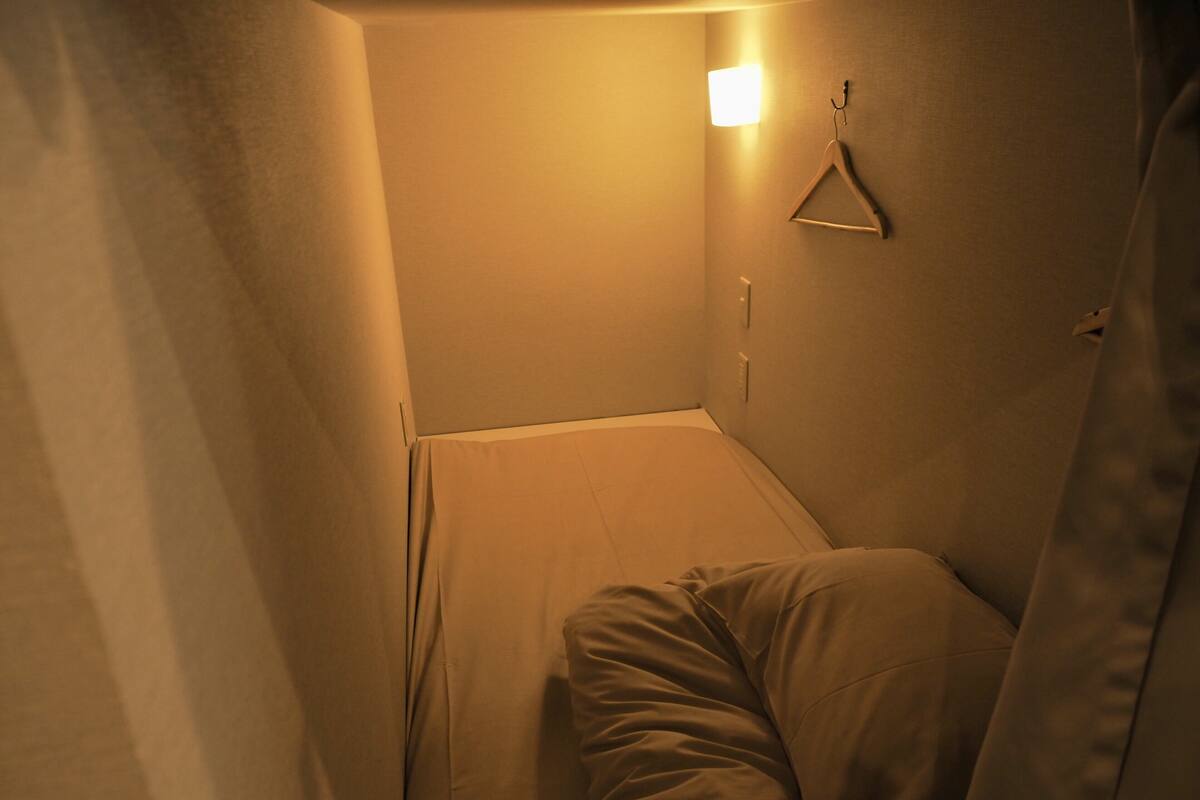 A narrow, dimly lit room with a single bed, a hanging light fixture, and a wall-mounted hanger.