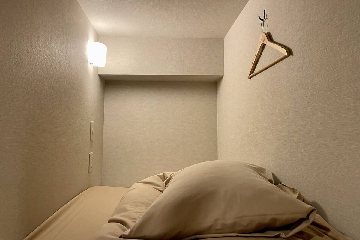 A small, dimly lit room with a single bed, a wooden hanger on the wall, and a light fixture above the bed.