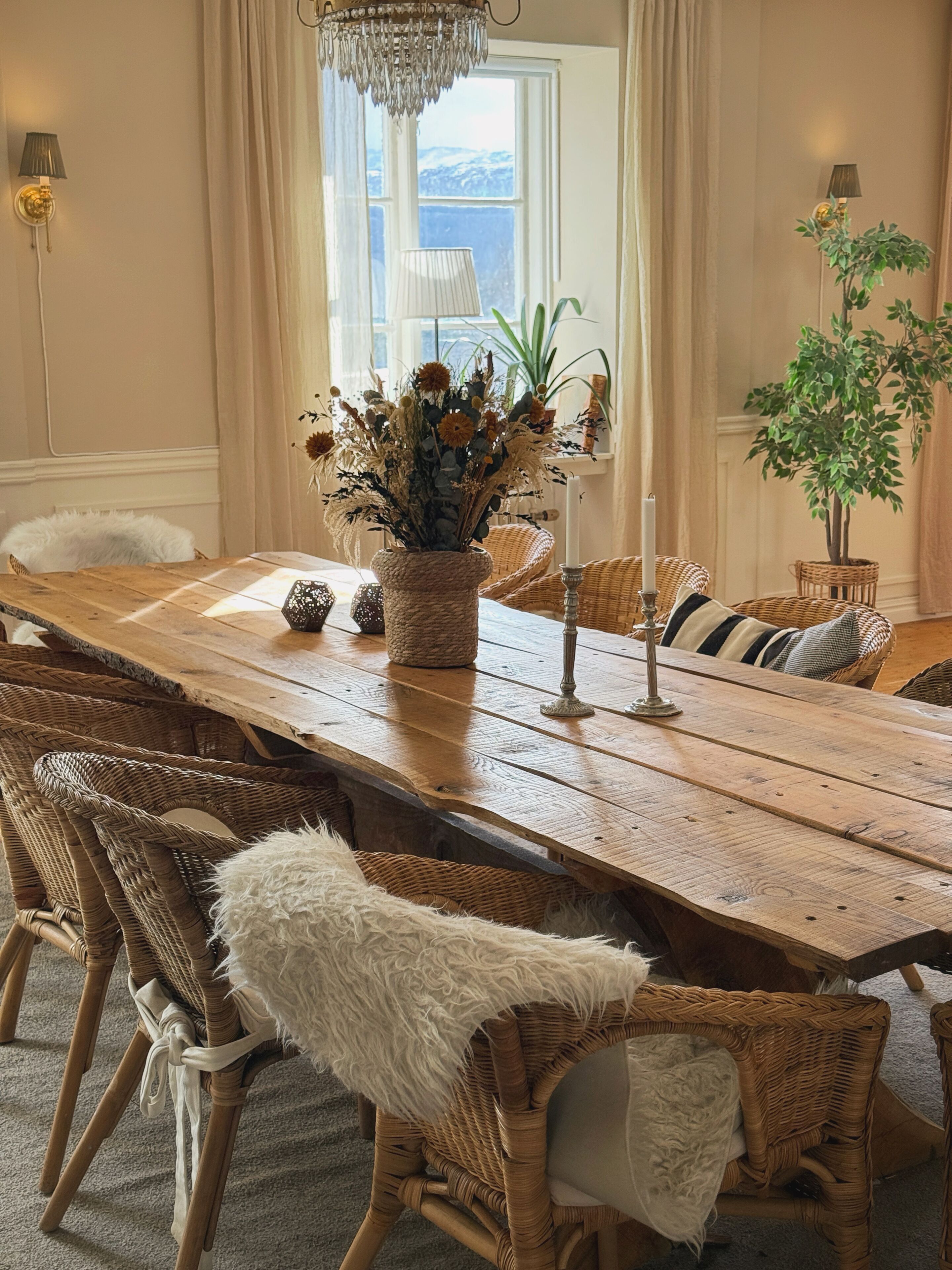 Un comedor con una mesa de madera, sillas de mimbre, una lámpara de araña y vistas al exterior a través de un ventanal.