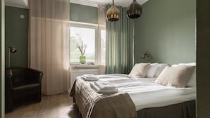 Un dormitorio con cama, una silla, un ventanal con cortinas y vistas al verdor exterior.
