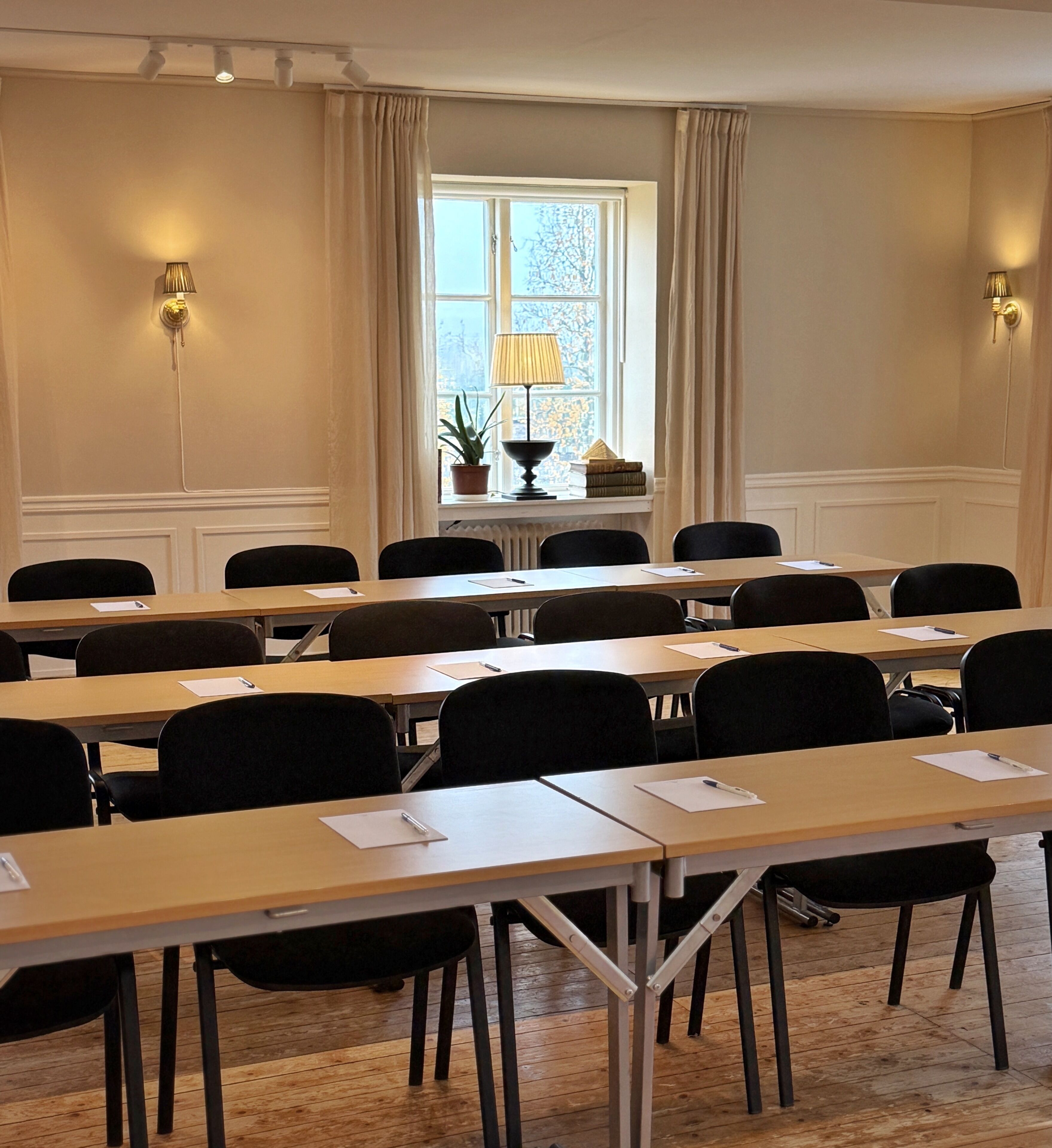 Ett konferensrum med långa bord, stolar, ett fönster med gardiner och väggmonterade lampor.