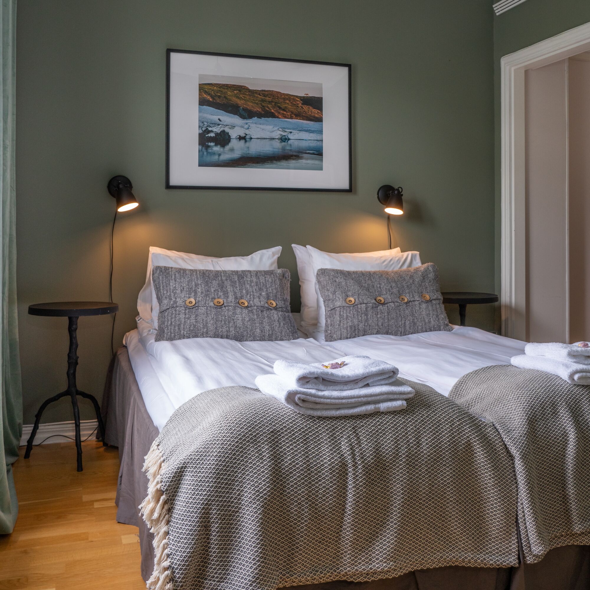 Una cama doble bien hecha con una colcha gris y blanca estampada, dos almohadas grises con botones de madera y un cuadro con paisaje sobre la cama.