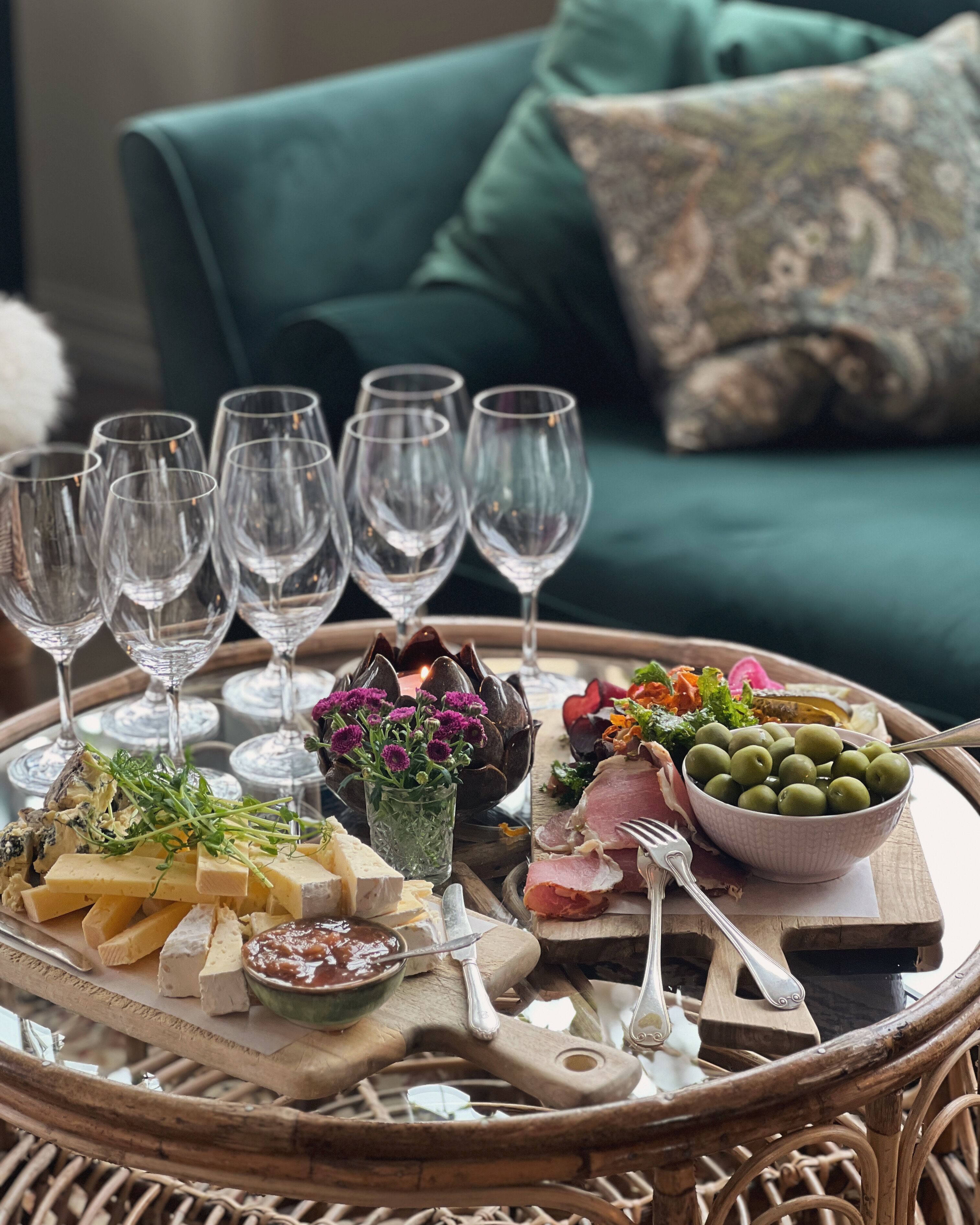 A wicker tray with various cheeses, olives, and a bowl of greens, accompanied by wine glasses and cutlery, set on a table in a room with a green sofa and patterned cushions.