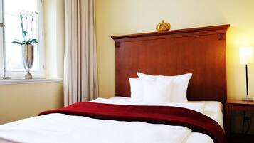 A hotel room with a wooden headboard, white bedding, a red throw blanket, a bedside lamp, and a window with curtains and a vase of flowers.