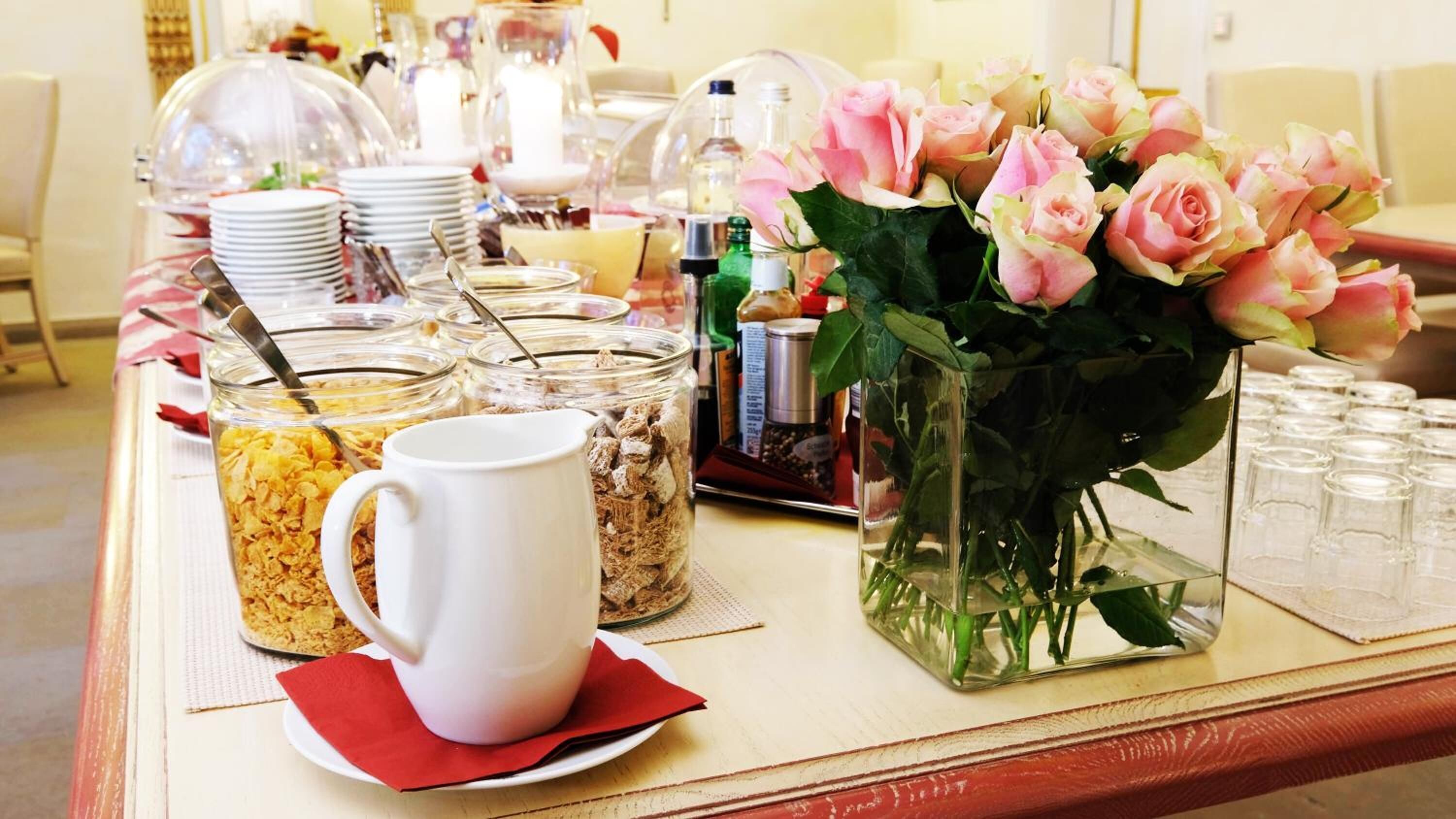 A breakfast buffet setup with various food items, a coffee mug, and a vase of pink roses.