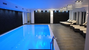 Ein Swimmingpool mit blau beleuchtetem Innenraum, einem Holzboden und Liegestühlen an der rechten Seite.