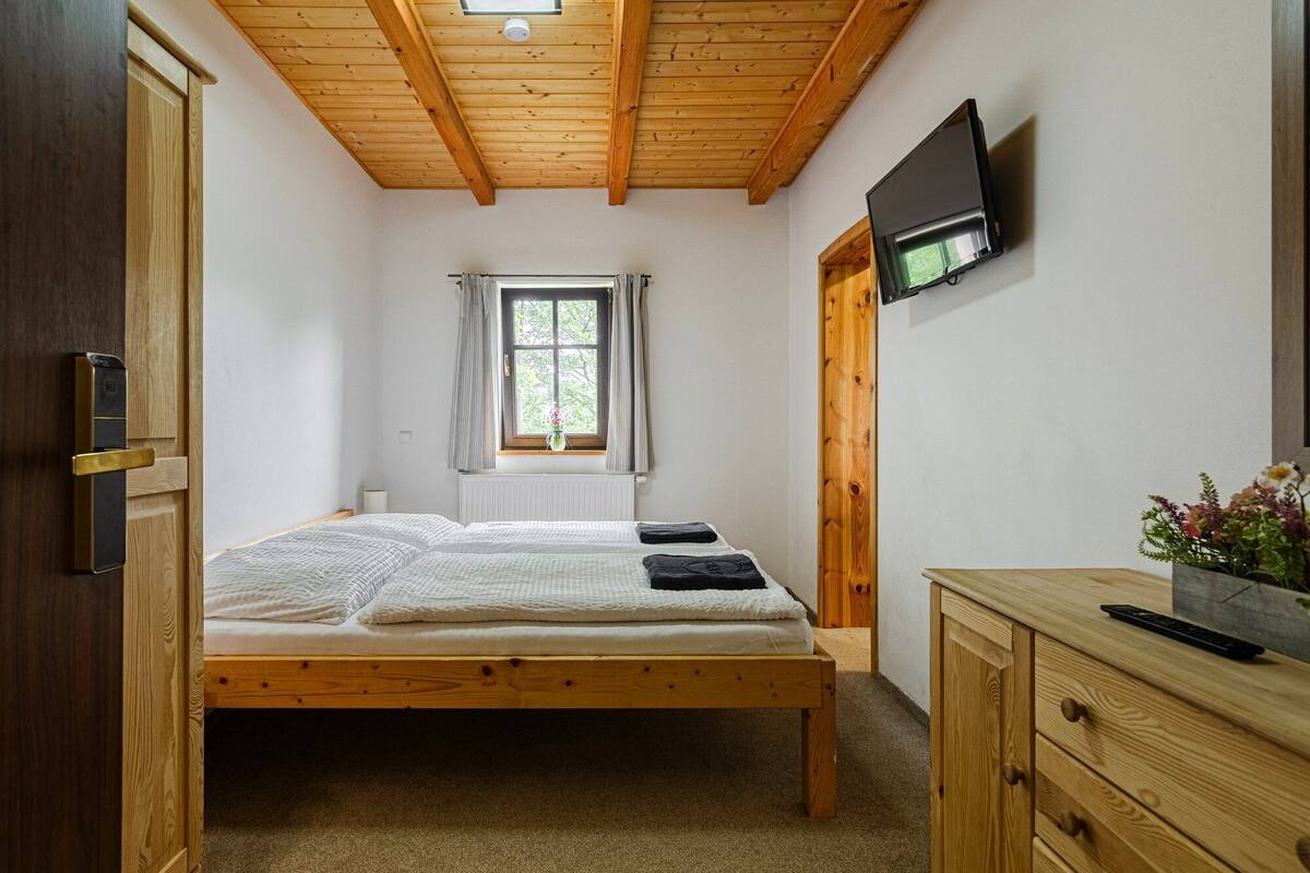 A bedroom with a wooden bed, a window with curtains, a TV mounted on the wall, and a wooden dresser.