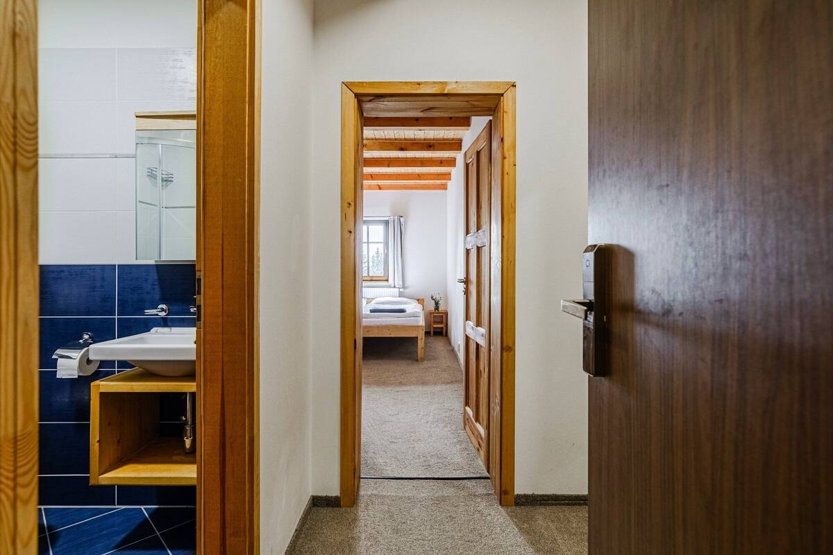 A hallway leading to a bedroom with a wooden door, a visible bathroom with a sink and mirror, and a window providing natural light.