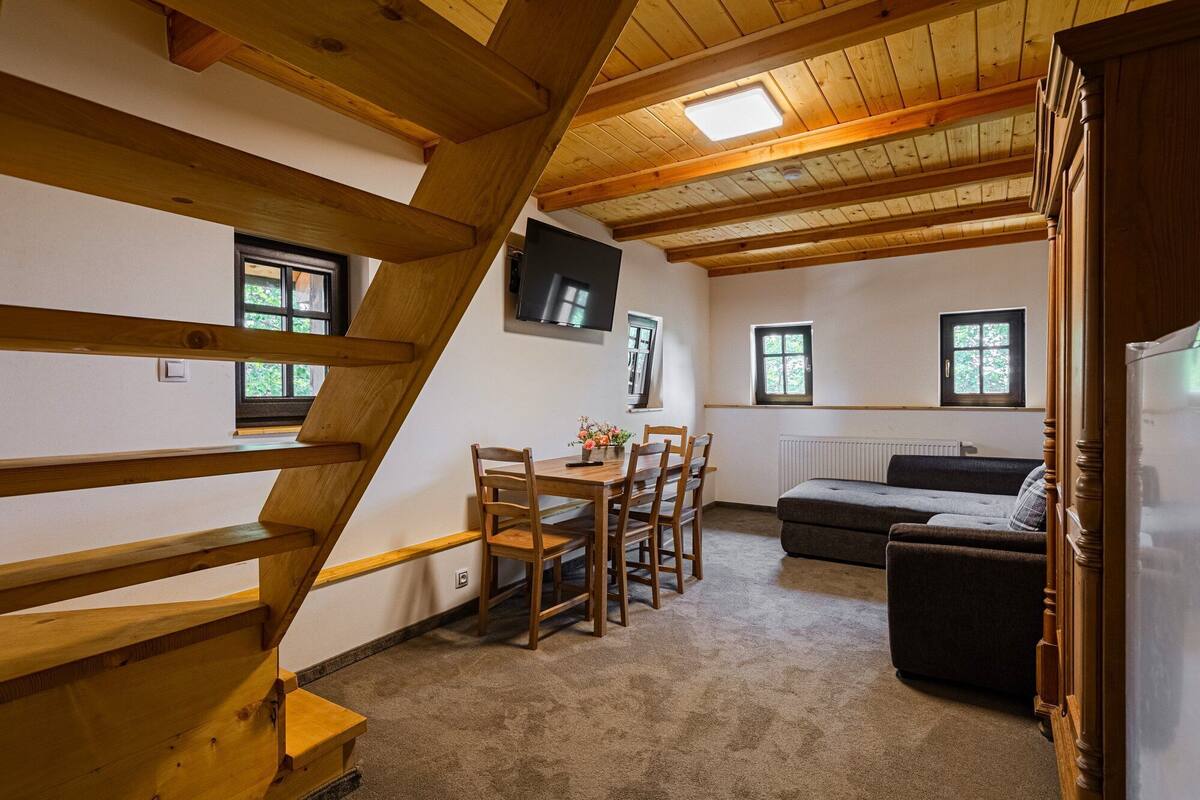 A cozy room with a wooden staircase, a dining area with a table and chairs, a sofa, and a television mounted on the wall.