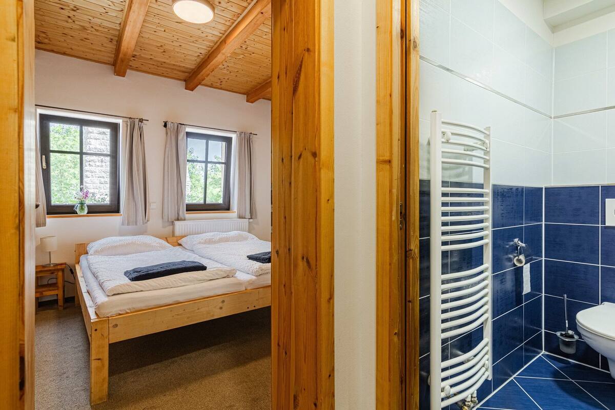 A bedroom with a wooden ceiling, two windows with curtains, a bed with white bedding, and a bathroom with blue tiles and a white bathtub.
