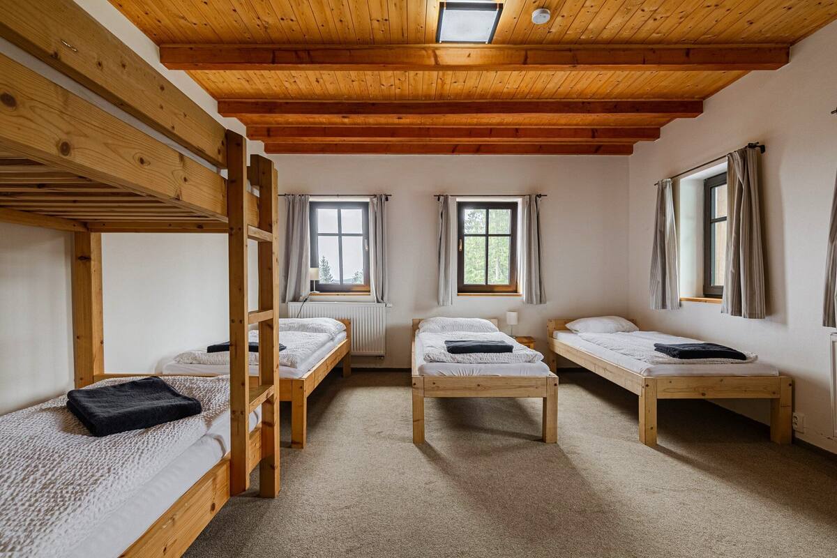 A dormitory room with bunk beds and two single beds, wooden beams on the ceiling, and windows with curtains.