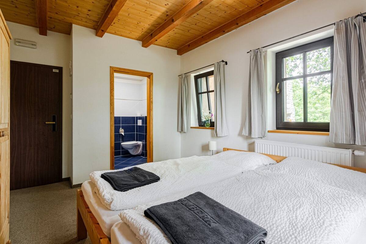 A bedroom with two beds, a window with curtains, a wooden ceiling, and a bathroom visible through an open door.