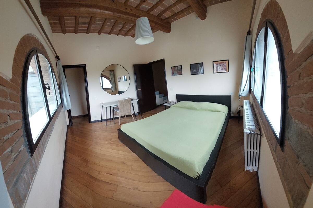 Un dormitorio con una cama grande, suelo de madera, paredes de ladrillo y ventanas amplias.