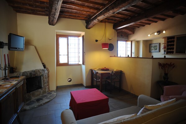 A cozy living room with a stone fireplace, a television mounted on the wall, a dining area with a table and chairs, and a red ottoman. - Agriturismo Il Poderaccio (Scandicci)