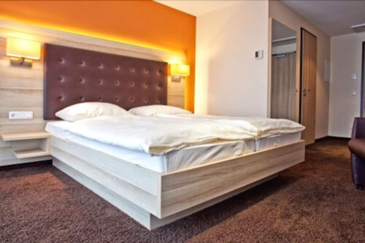 A hotel room with a large bed, a headboard with a light fixture, a wooden nightstand, and a carpeted floor.