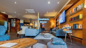 A modern, well-lit hotel lobby with a seating area, a wooden coffee table, and a bookshelf with books and a television screen. - Bacchus Hotel (Bensheim)