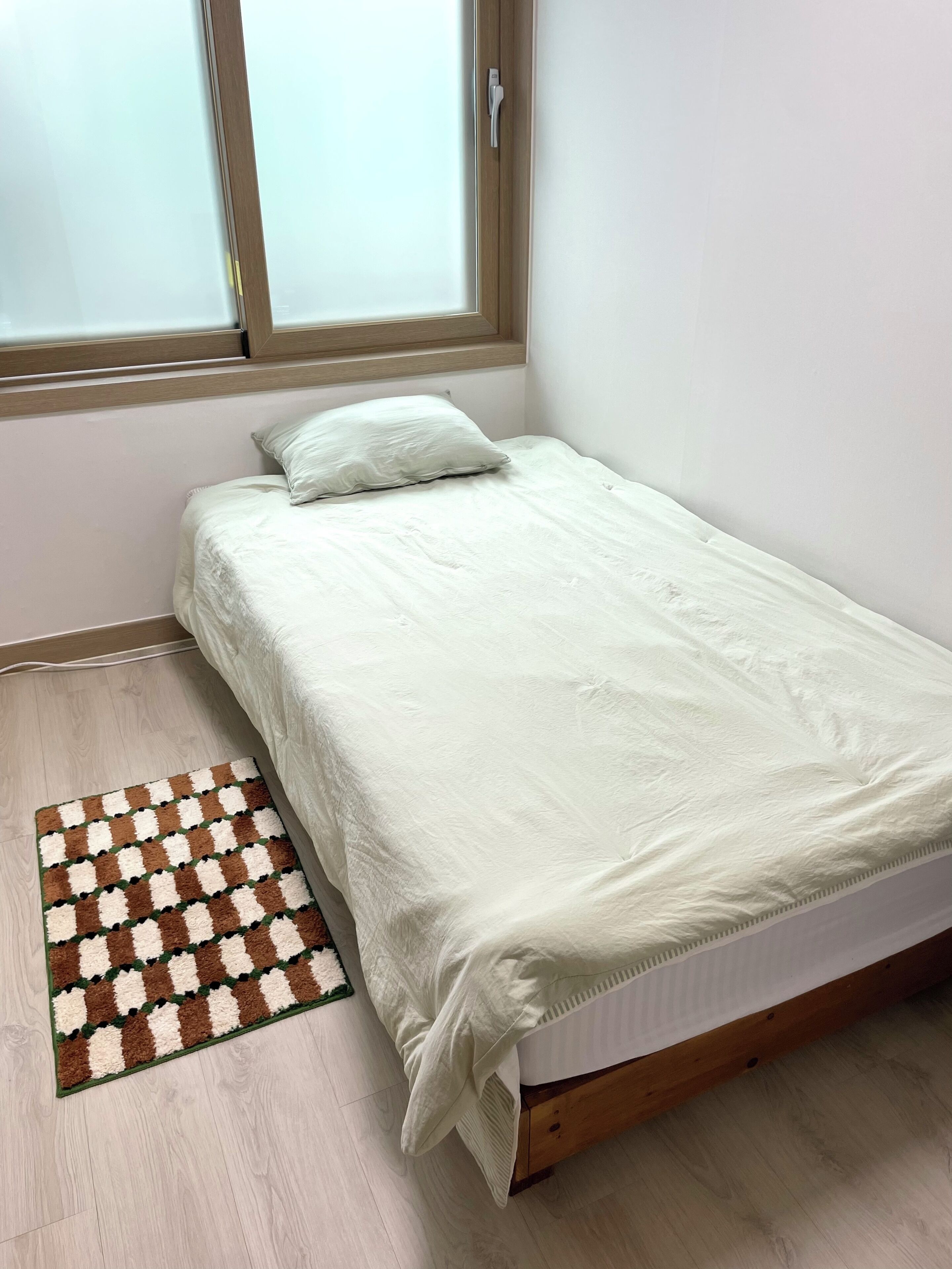 A small, neatly made bed with a white mattress and a matching pillow, positioned next to a window with a wooden frame.