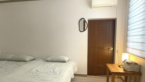 Un dormitorio con cama, una mesita de noche con lámpara, un pequeño mueble de madera y un cubo de basura blanco.