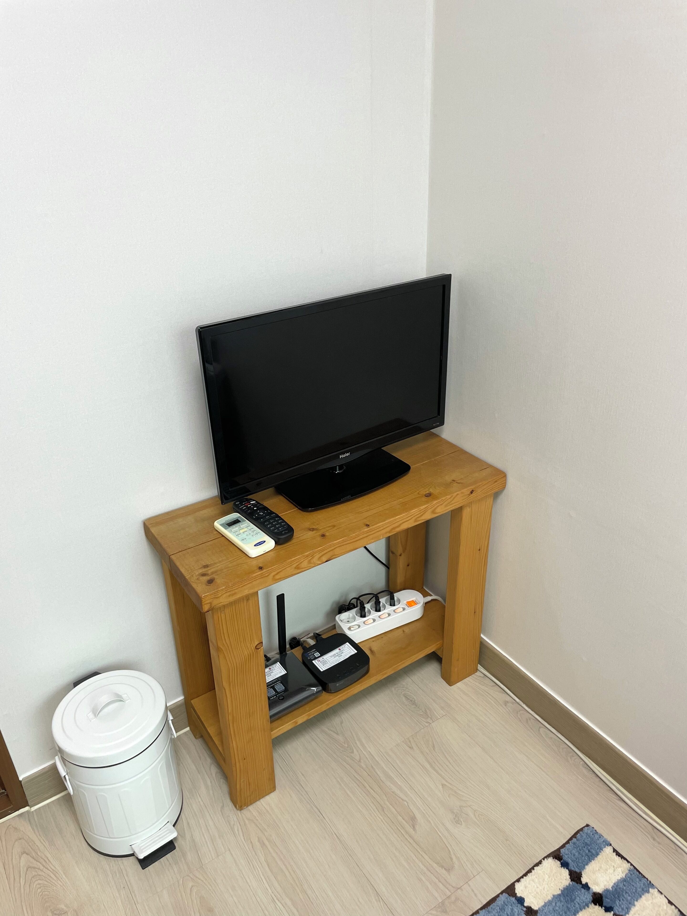 A television on a wooden stand with a remote, a white trash bin, and a power strip with cables.