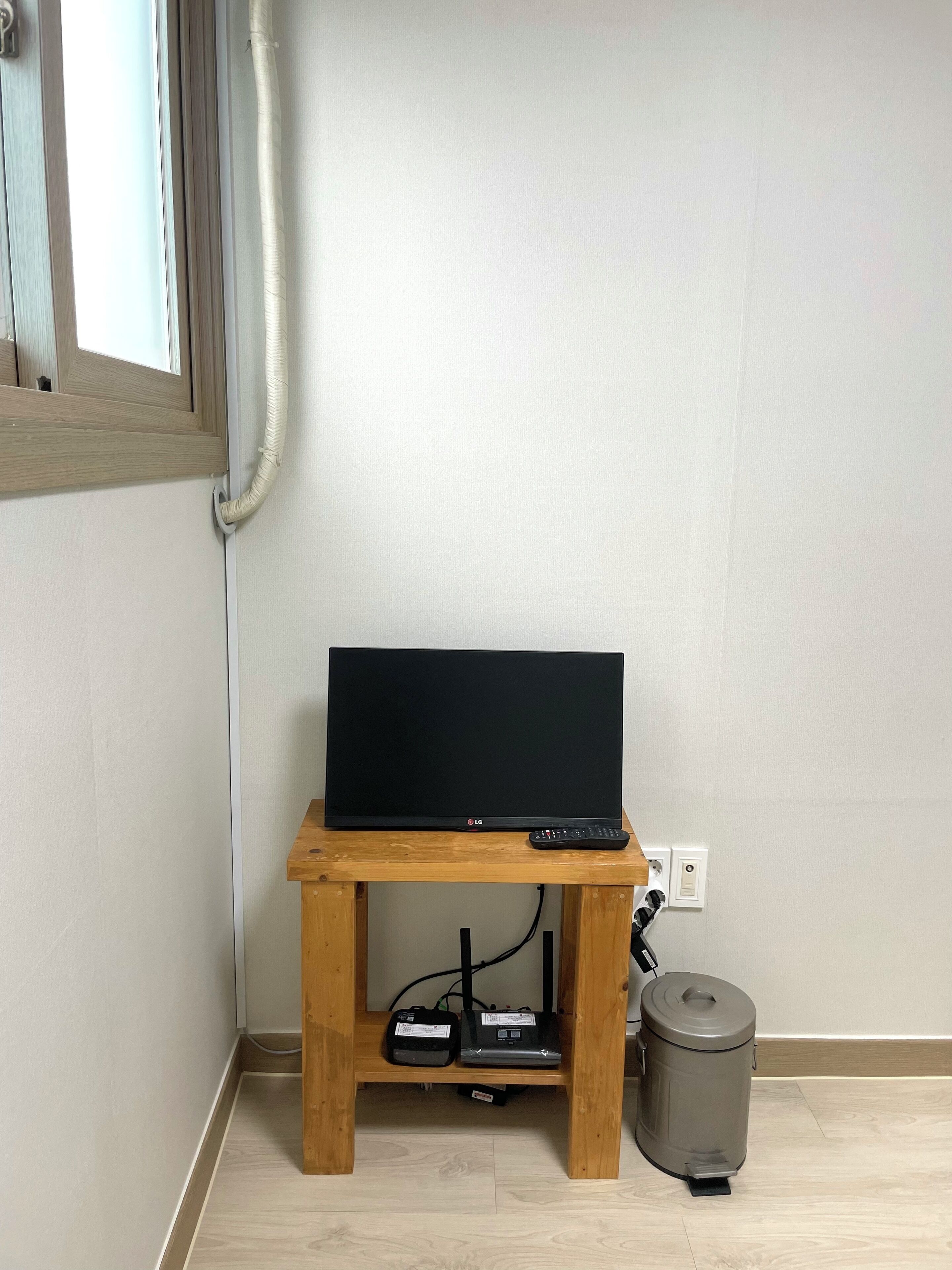 A television on a wooden stand with a remote control, a trash can, and a power outlet on the wall.