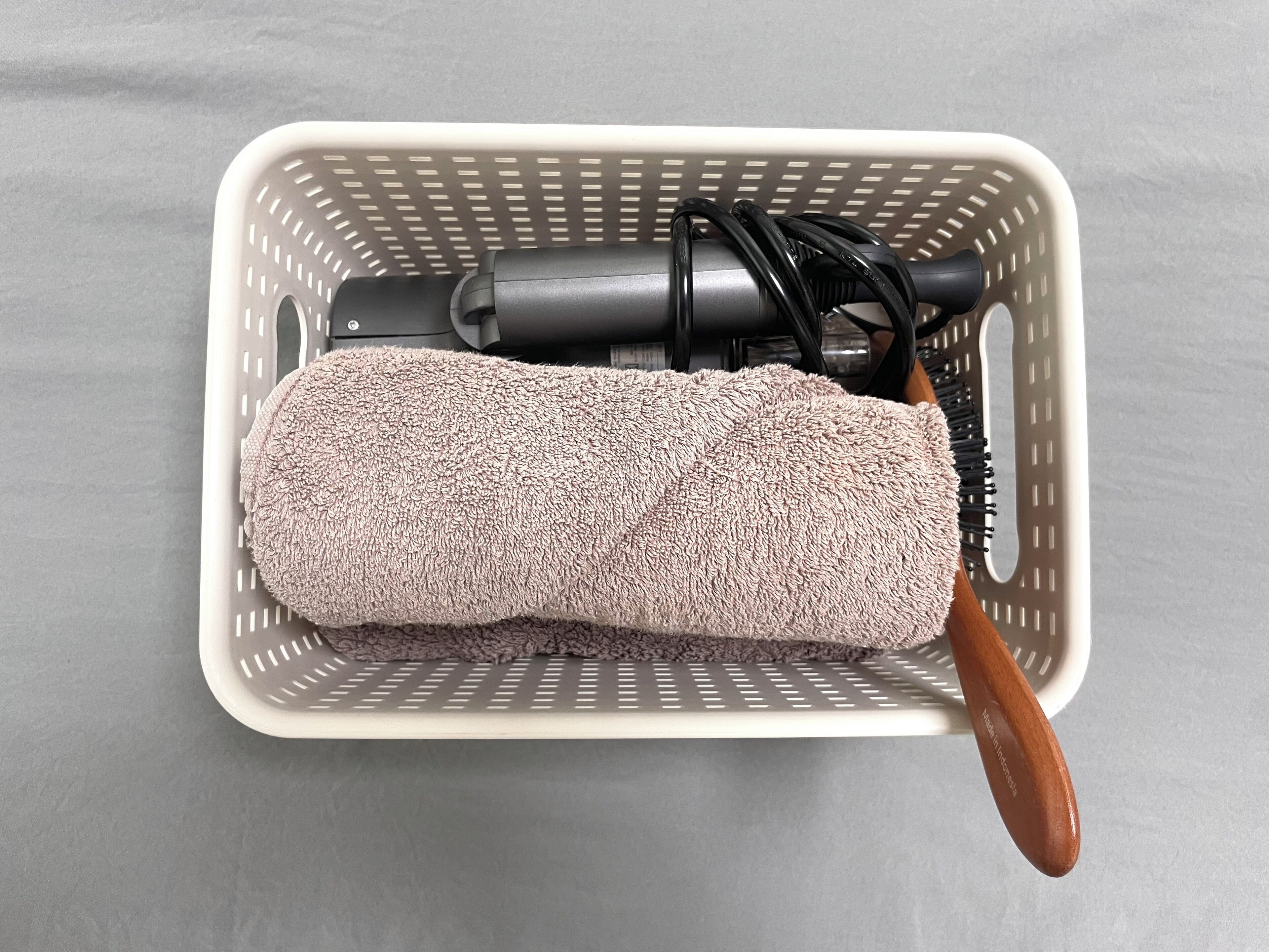 Una cesta blanca con una toalla enrollada, un cepillo negro para el cabello y una goma para el pelo negra sobre una superficie gris.